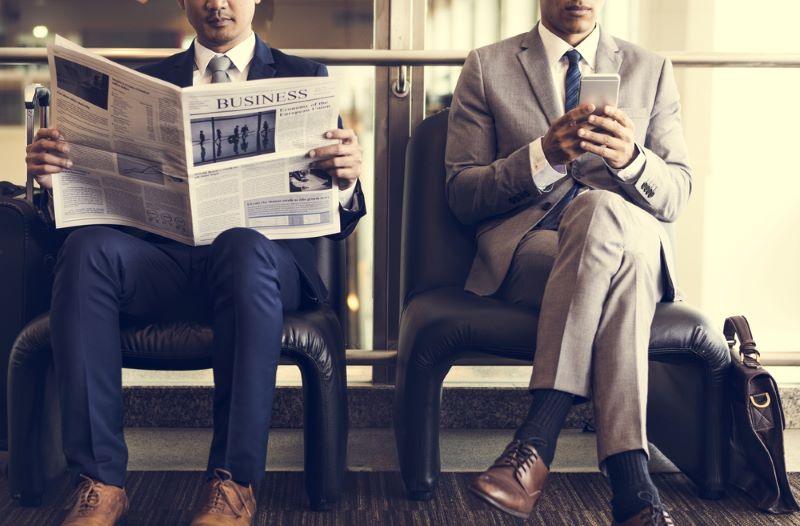 Deux hommes en costume patientent : l'un d'entre eux lit le journal 