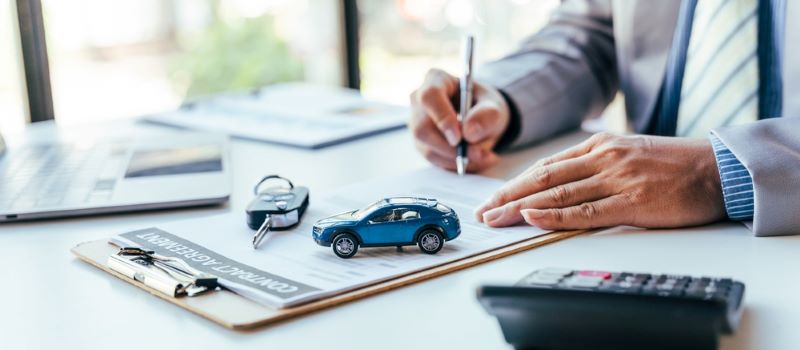 Un homme signe un contrat d'assurance posé sur un bureau avec, devant lui, un petit porte-clés voiture