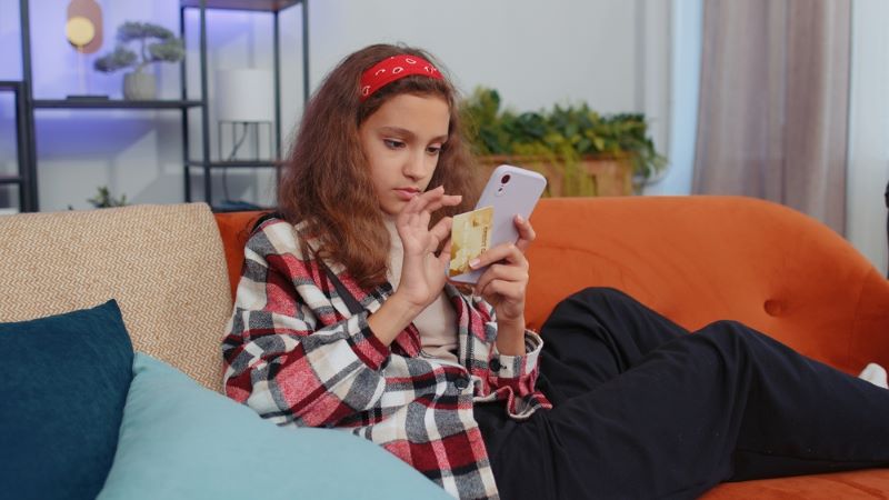 Une très jeune femme assise sur son canapé manipule son portable et sa carte Axa